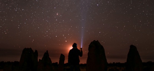 A silhouette of a person standing among large rocks shining a small light on a sky full of stars