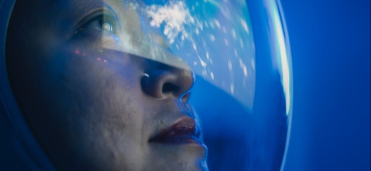 A close-up of a woman’s face looking out from her space helmet at the universe
